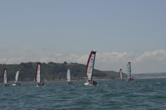 Sailboats glide on the ocean near a hilly coastline under a clear sky, in Brittany, France