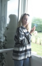 A woman with a striped shirt stands by a window, holding a cigarette, with natural light