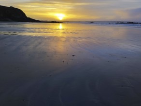 A serene sunset over a beach with calm ocean waters and rocks silhouetted against a golden sky