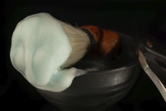 Shaving brush with teal lathered foam and straight razor in a dark bowl for men's grooming