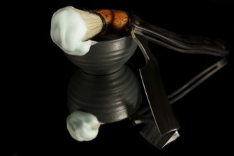 Classic straight razor, bowl, and brush with foam reflecting on a dark surface