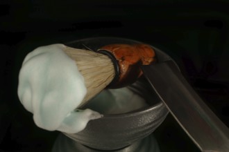 Shaving brush with cream and straight razor resting in a bowl on black background