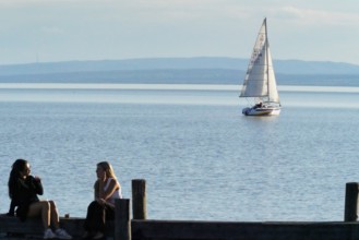 Lake Neusiedl, Burgenland, Austria, Europe, Two woman sit on the jetty while a sailing boat passes