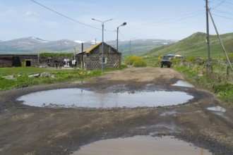A muddy road with large puddles leads through a village with mountains in the background, Zorakert,