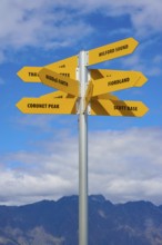Yellow signposts with various destinations against a backdrop of blue sky and mountains,