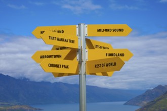 Yellow signpost with several signs in front of a mountain backdrop under a cloudy sky, Queenstown,