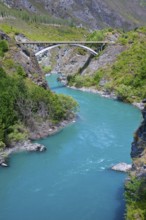 A turquoise-coloured river flows through a green valley with a bridge in the background, Kawarau