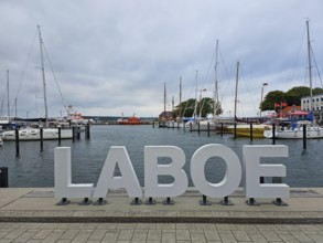 Harbour with large LABOE lettering in the foreground, surrounded by boats and a cloudy sky, Laboe,