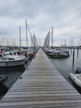 A long jetty runs through a harbour, surrounded by boats and masts on a calm, cloudy day, Laboe,