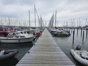 A wooden walkway leads through a harbour, lined with boats and masts under a cloudy sky, Laboe,