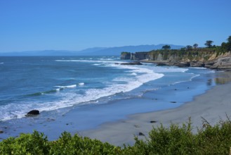 A peaceful coastal landscape with blue waves crashing against sand and cliffs under a clear blue
