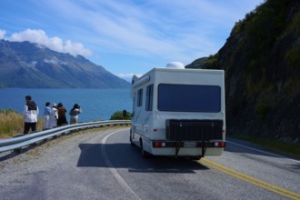 People enjoying travelling and a motorhome driving on a road along a lake surrounded by mountains,