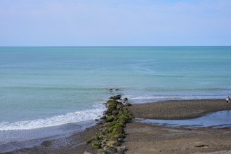 View of the calm sea with rocks leading into the water and a lone walker on the beach, Mana Bay,