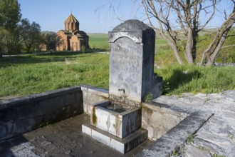 Stone fountain with engraving in front of a rural church in a green landscape, original spring,