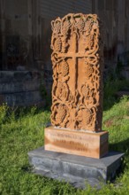 A detailed engraved red gravestone with a cross motif stands on a grassy area in the sunlight,