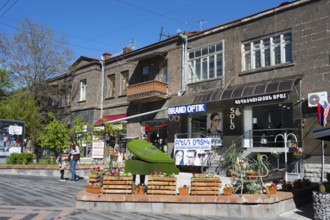 Pedestrian zone with optical shop and advertising signs, flanked by concrete buildings and trees,