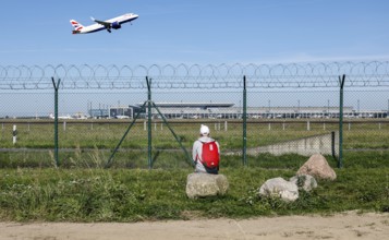 A man sits on a stone and watches the take-off of a British Airways plane through a barbed wire