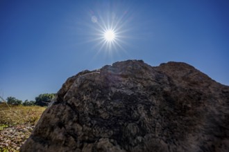 A large stone stands in the foreground under a bright blue sky. The sun shines brightly and creates