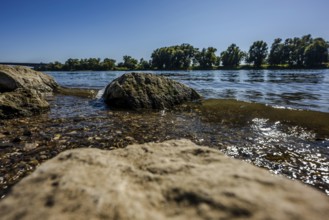 The scene shows a calm river with large stones lying on its banks. The water glistens in the sun,