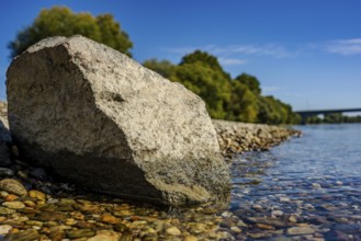 A large stone lies on the bank of a calm river. The water is clear and reflects the blue sky. Trees
