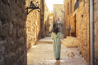 Woman in traditional abaya walking with cat through narrow stone alley in historic city of Mardin