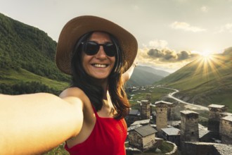 Smiling woman in hat and red dress taking selfie at sunset with medieval towers of Ushguli Georgia