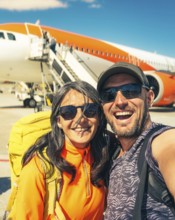 Cheerful caucasian couple taking selfie in front of passenger airplane before boarding flight