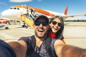Cheerful caucasian couple taking selfie in front of passenger airplane before boarding flight