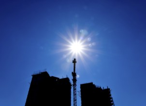 Tower crane under construction between the two high-rise buildings against the blue sky and sun.