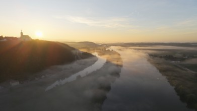 In the early morning, thick fog lies over the river. The sun slowly rises and illuminates the