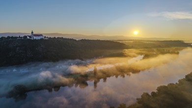 The sunrise illuminates the landscape over the calm Danube. A light mist floats gently over the