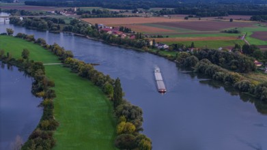 A cargo ship sails quietly on the Danube. The surroundings are green with fields and trees. The day