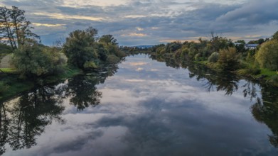 A calm river landscape at dusk shows trees and clouds in the reflecting water surface. Nature