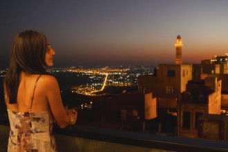 Tourist woman on terrace restaurant balcony watching illuminated skyline of Mardin with glowing