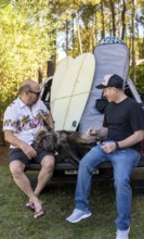 Two male friends talking sitting on a truck with their dog and surfboards
