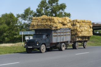 A lorry loaded with bales of straw drives along a country road between green trees, Armenia