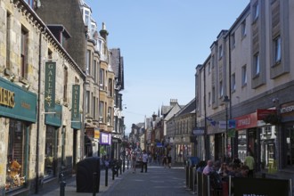 Busy shopping street with people and traditional buildings under a blue sky, Fort William,