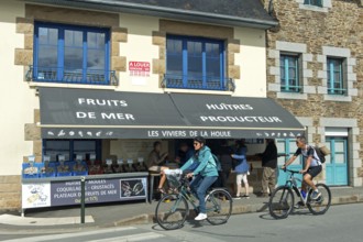 Cyclists on the Quai John Kennedy pass a shop selling seafood such as oysters and mussels, Cancale,