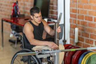 Young man living with a disability sitting in a wheelchair while adjusting a barbell rack during a
