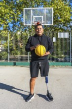 Man with prosthetic leg stands on outdoor basketball court holding a yellow ball, exuding