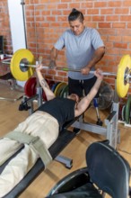Young man with a prosthetic leg performing bench press exercises at the gym, receiving support and