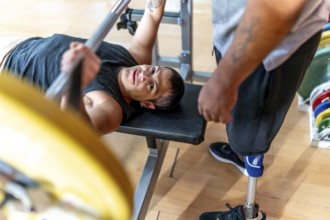 Disabled man performing a bench press with a barbell while a friend with a prosthetic leg stands