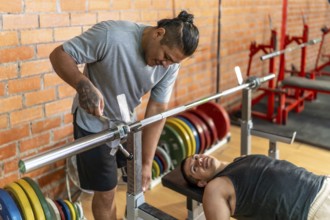 Two diverse men, one with a prosthetic limb assisting the other who is laying on a weight bench,