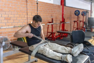 Young man fitting a lifting strap onto his prosthetic leg before a gym workout, focused and