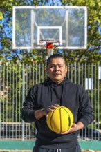 Indigenous man standing on an outdoor basketball court holding a bright yellow basketball, looking