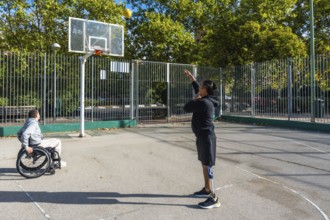 Two athletes playing basketball on an outdoor court. One sitting in a wheelchair and the other