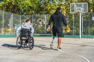 Man with prosthetic leg dribbling basketball on outdoor court while friend in a wheelchair watches,