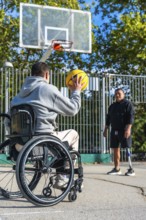 Man in a wheelchair aiming to shoot a basketball while his male friend, who uses a prosthetic leg,