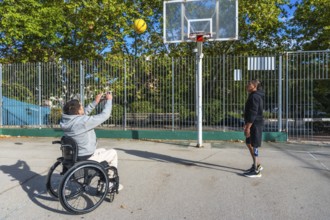 Two friends with disabilities play basketball outdoors, one shoots from a wheelchair while the