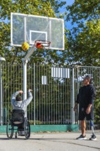 Two men play outdoor basketball, one shooting from a sports wheelchair while the other stands with